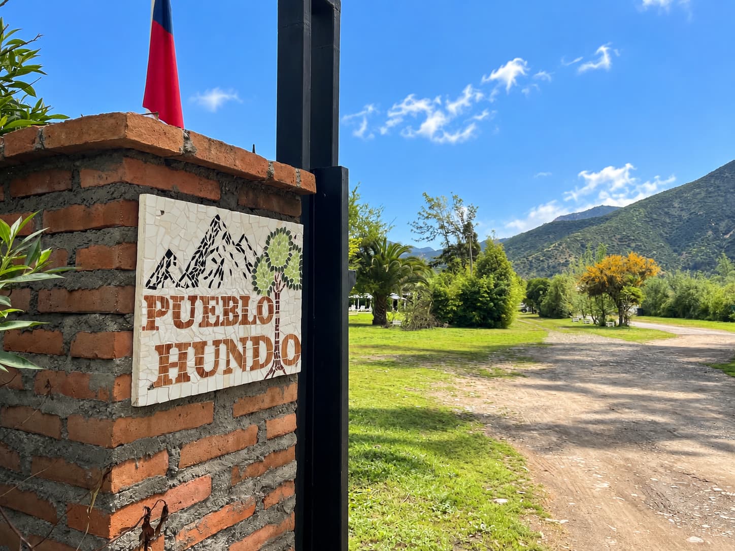 Letrero de mosaico de Pueblo Hundio en un pilar de ladrillo con la bandera chilena, junto al camino de entrada y el cerro