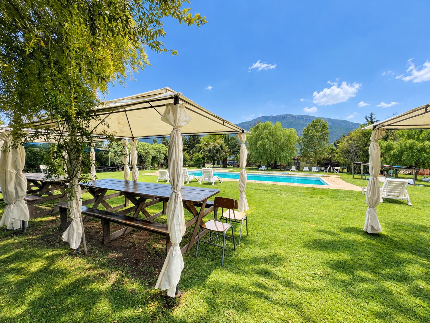 Toldo blanco con mesa de madera junto a la piscina, con reposeras y el cerro de fondo bajo el cielo despejado