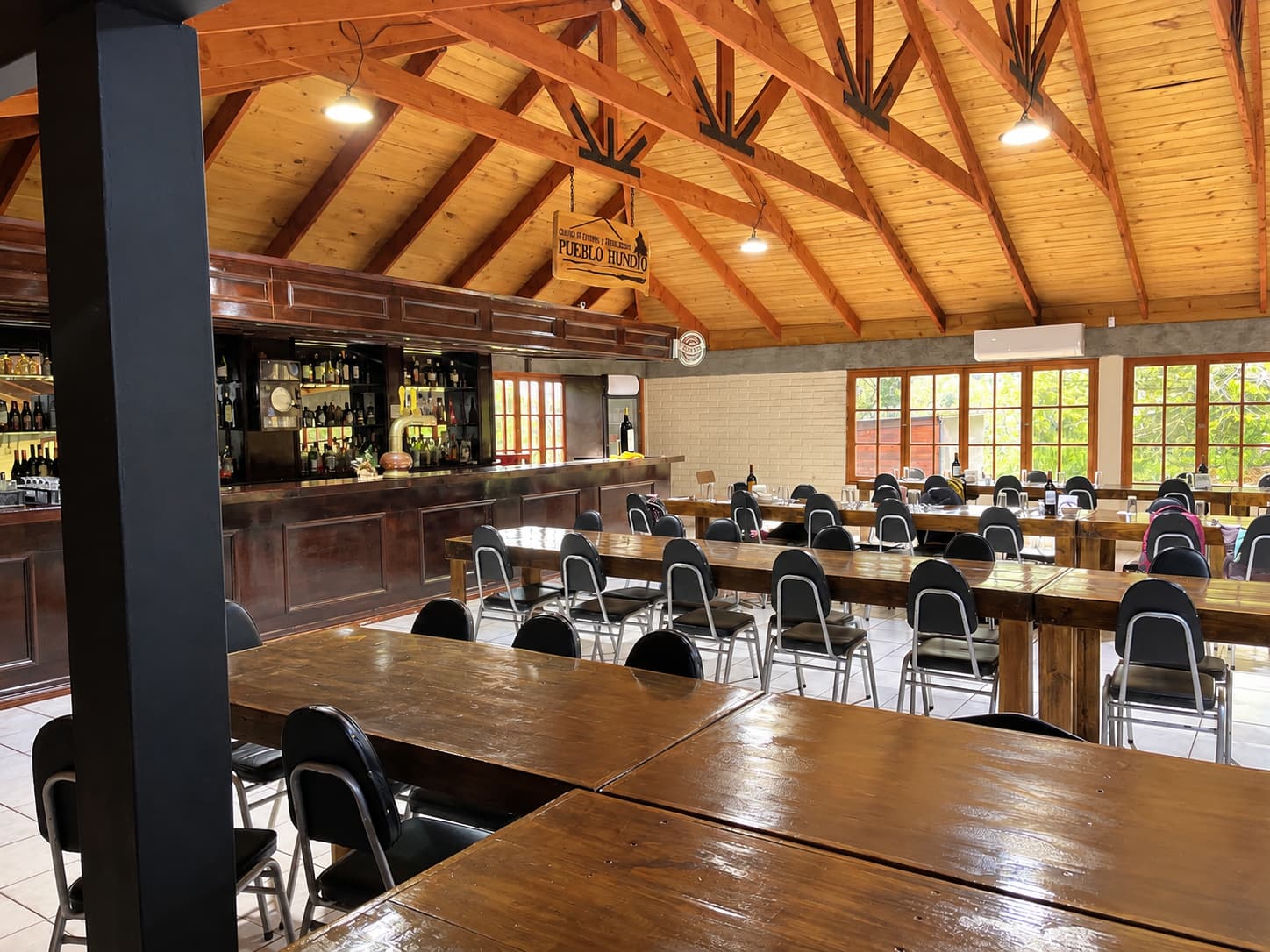 Salón con techo de vigas de madera, letrero de Pueblo Hundio, barra de madera y mesas largas con sillas negras
