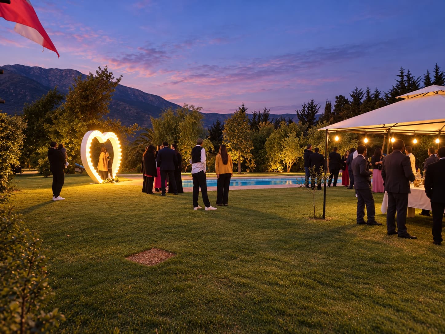 Invitados de gala en el jardín al atardecer, con un corazón de luces, la piscina y un toldo con guirnaldas bajo el cielo morado
