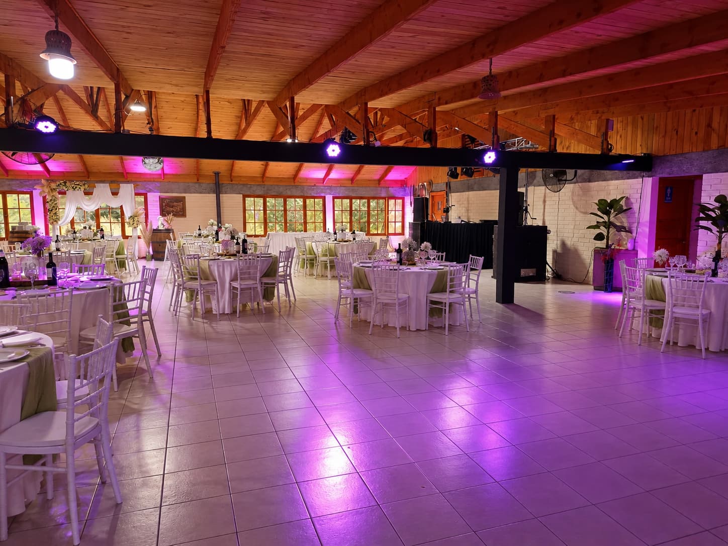 Salón montado para un matrimonio con mesas redondas, sillas blancas y luces moradas