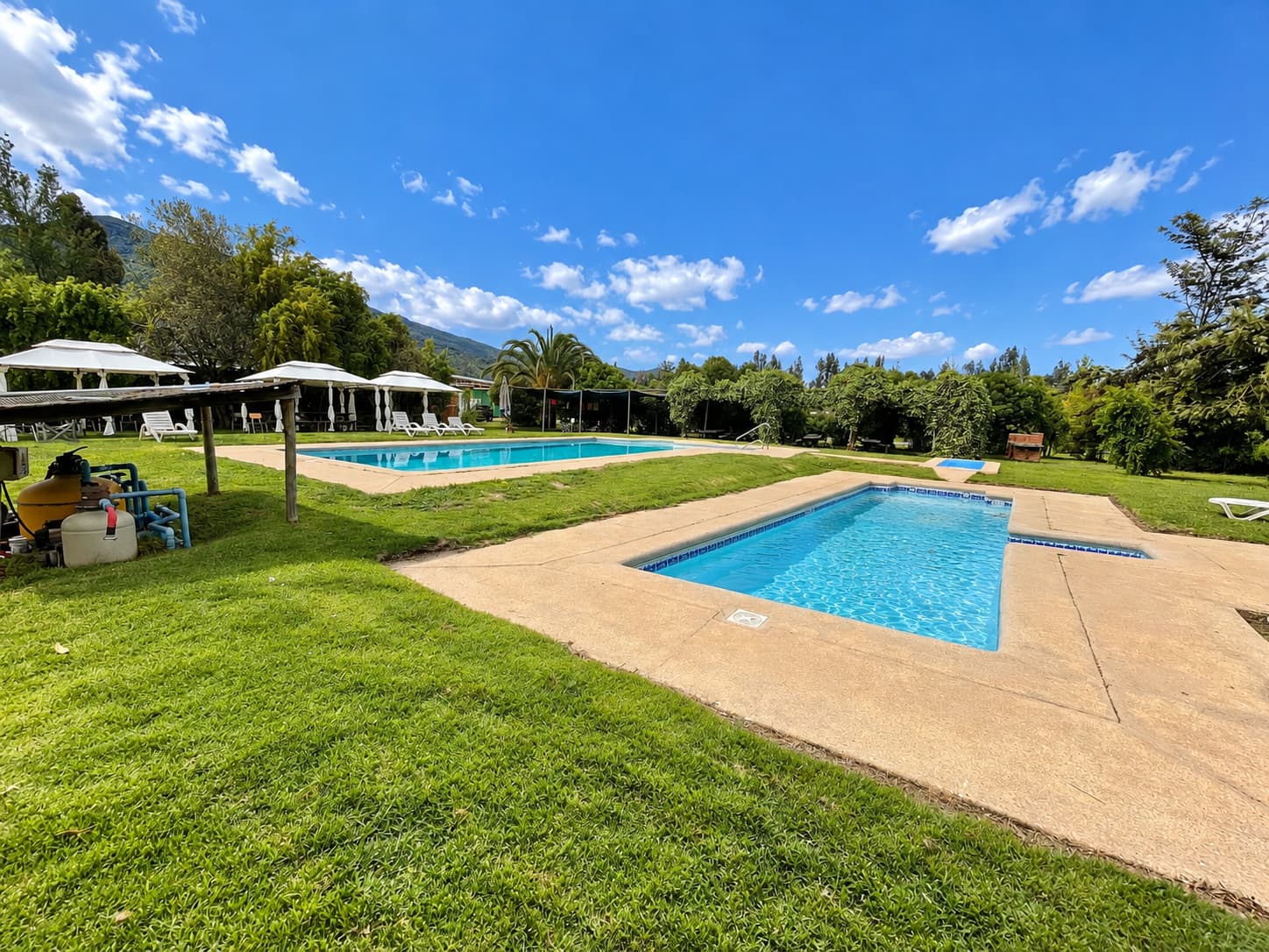Piscina chica en primer plano y piscina grande detrás, rodeadas de pasto, con toldos blancos y cerros al fondo