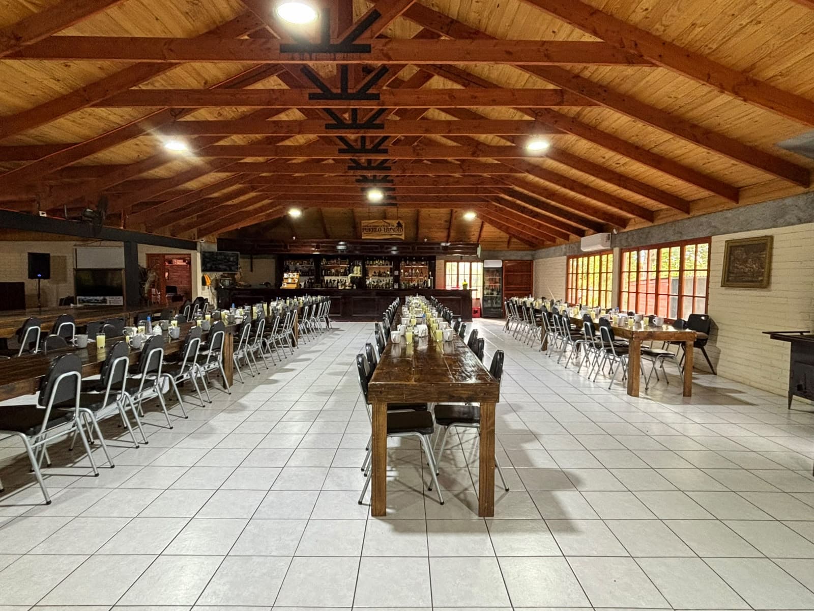 Salón completo con tres hileras de mesas largas preparadas para desayuno y la barra al fondo