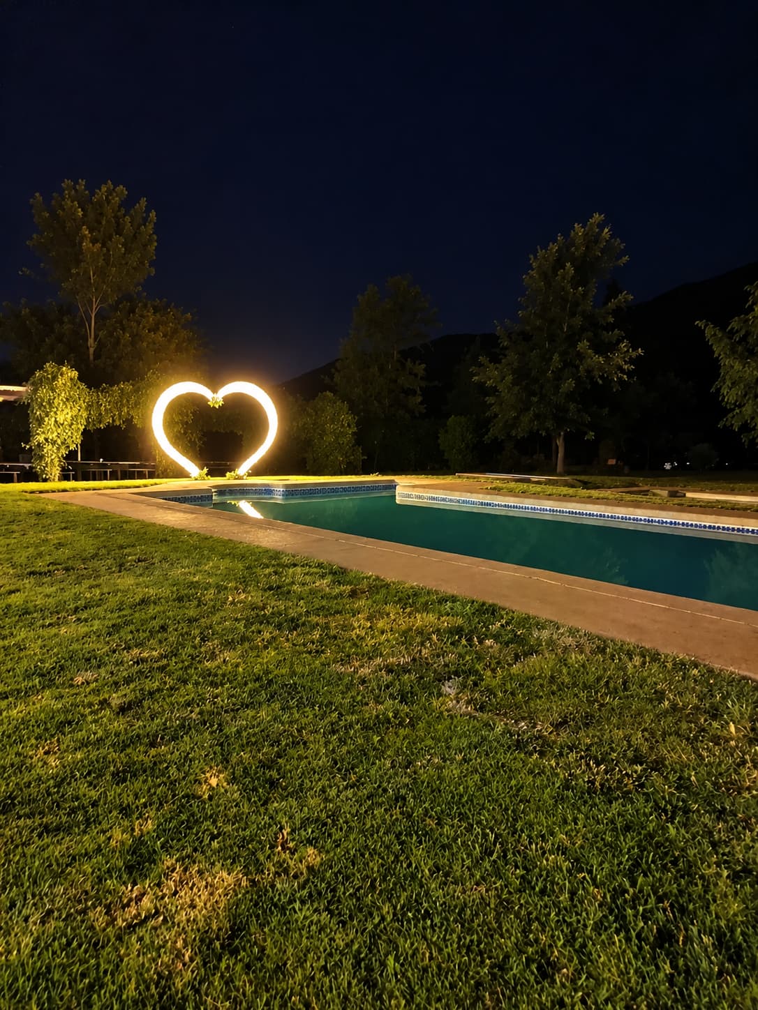 Evento matrimonio: corazón de luces encendido de noche junto a la piscina, con árboles y cerros oscuros detrás