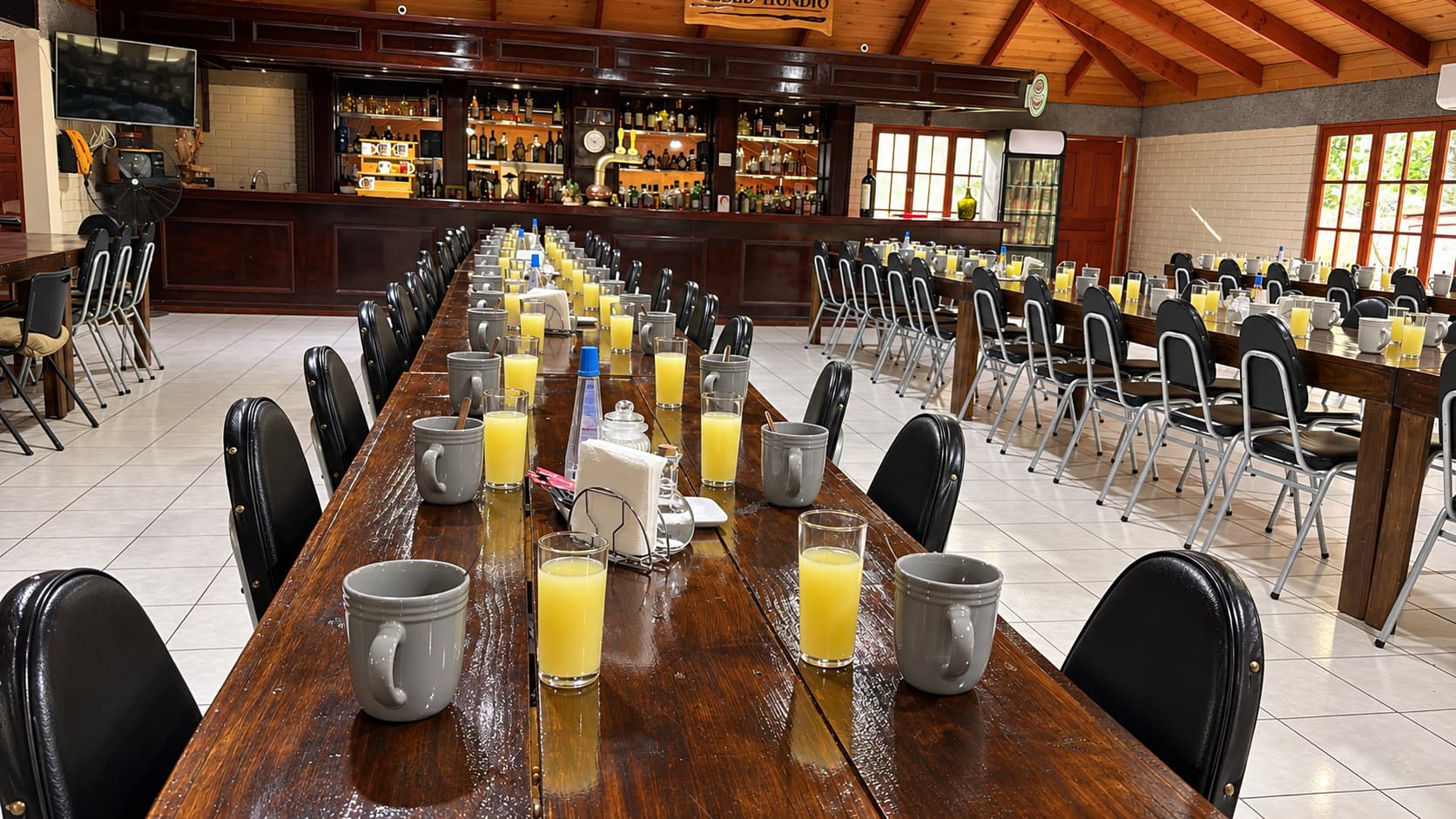 Mesa larga de madera con tazas grises y vasos de jugo, con la barra de licores al fondo
