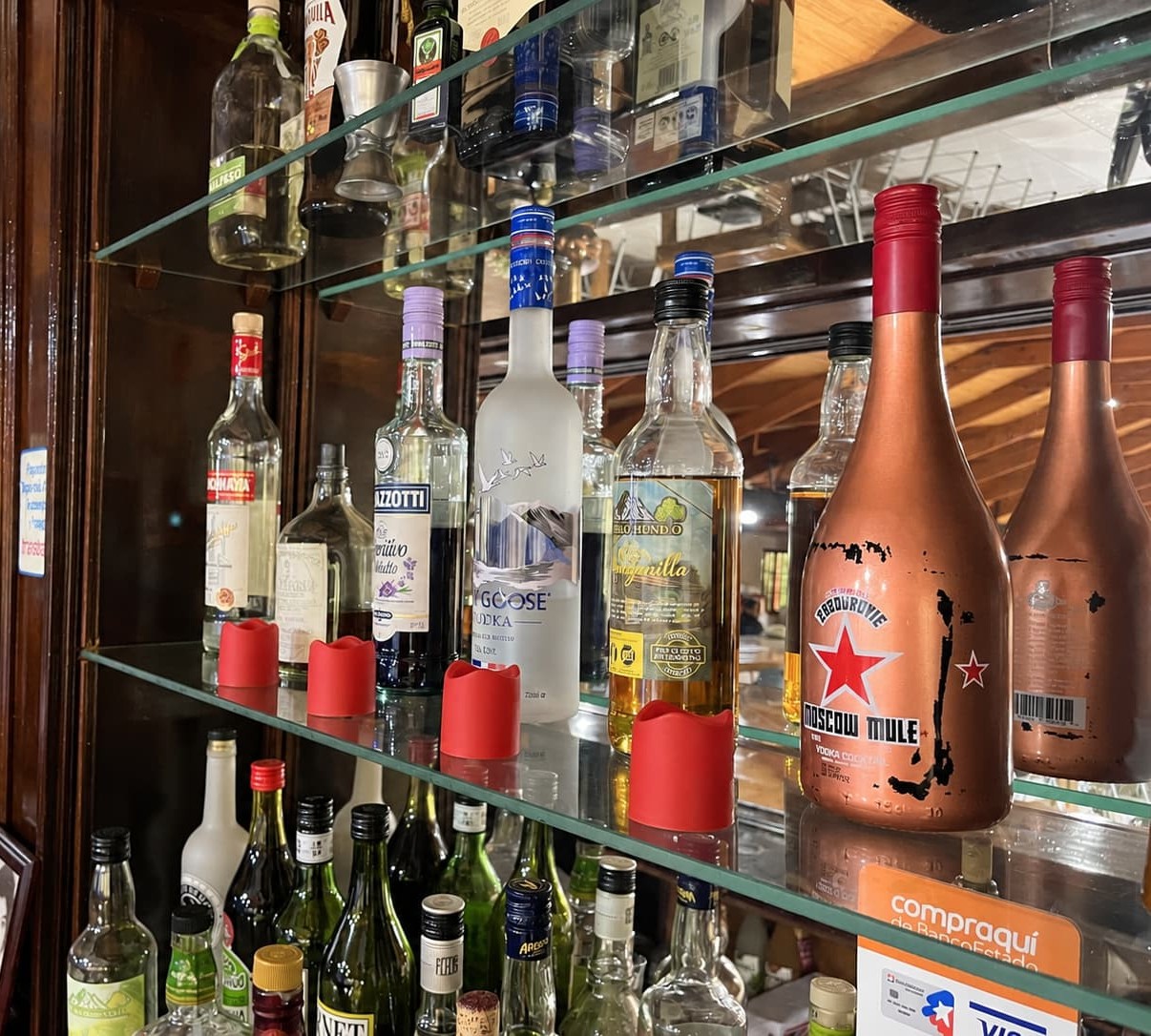 Repisas de vidrio del bar con botellas de vodka, Moscow Mule, licores y vinos, junto a velas rojas y un mueble de madera