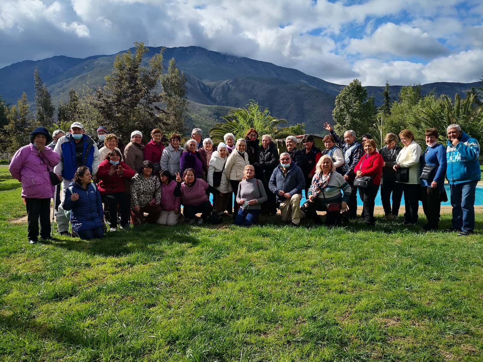 Grupo de visitantes posando en el pasto frente a la piscina, con la cordillera y nubes de fondo