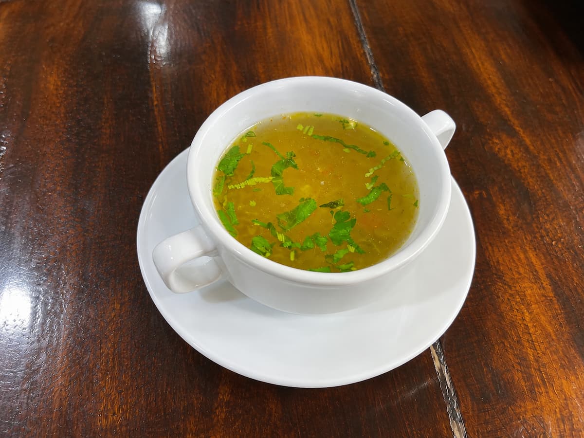 Consomé de ave con cilantro picado en taza blanca con asas, sobre platillo y mesa de madera