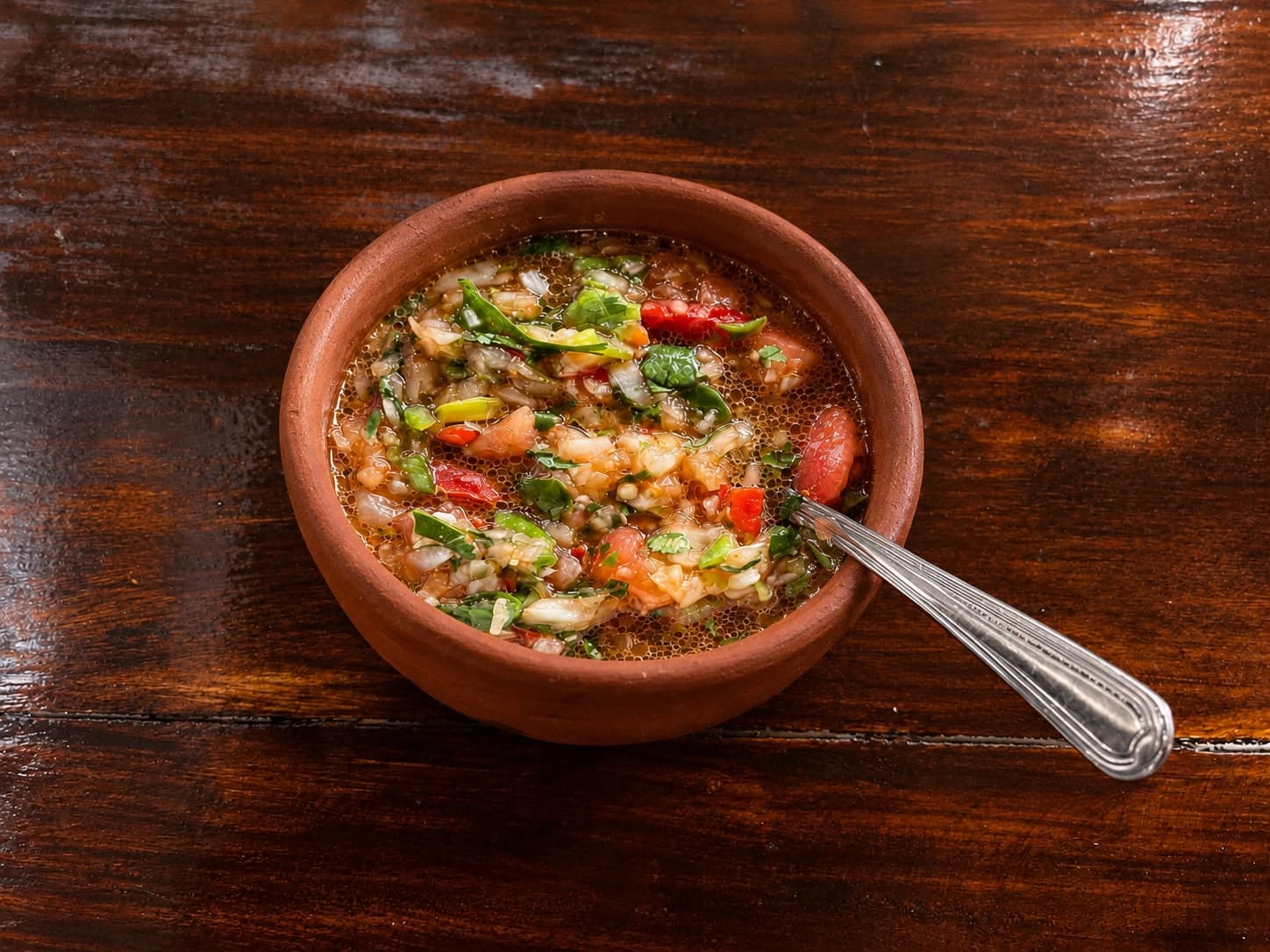 Pebre casero de tomate, cebolla y cilantro en un tazón de greda sobre mesa de madera, con una cuchara