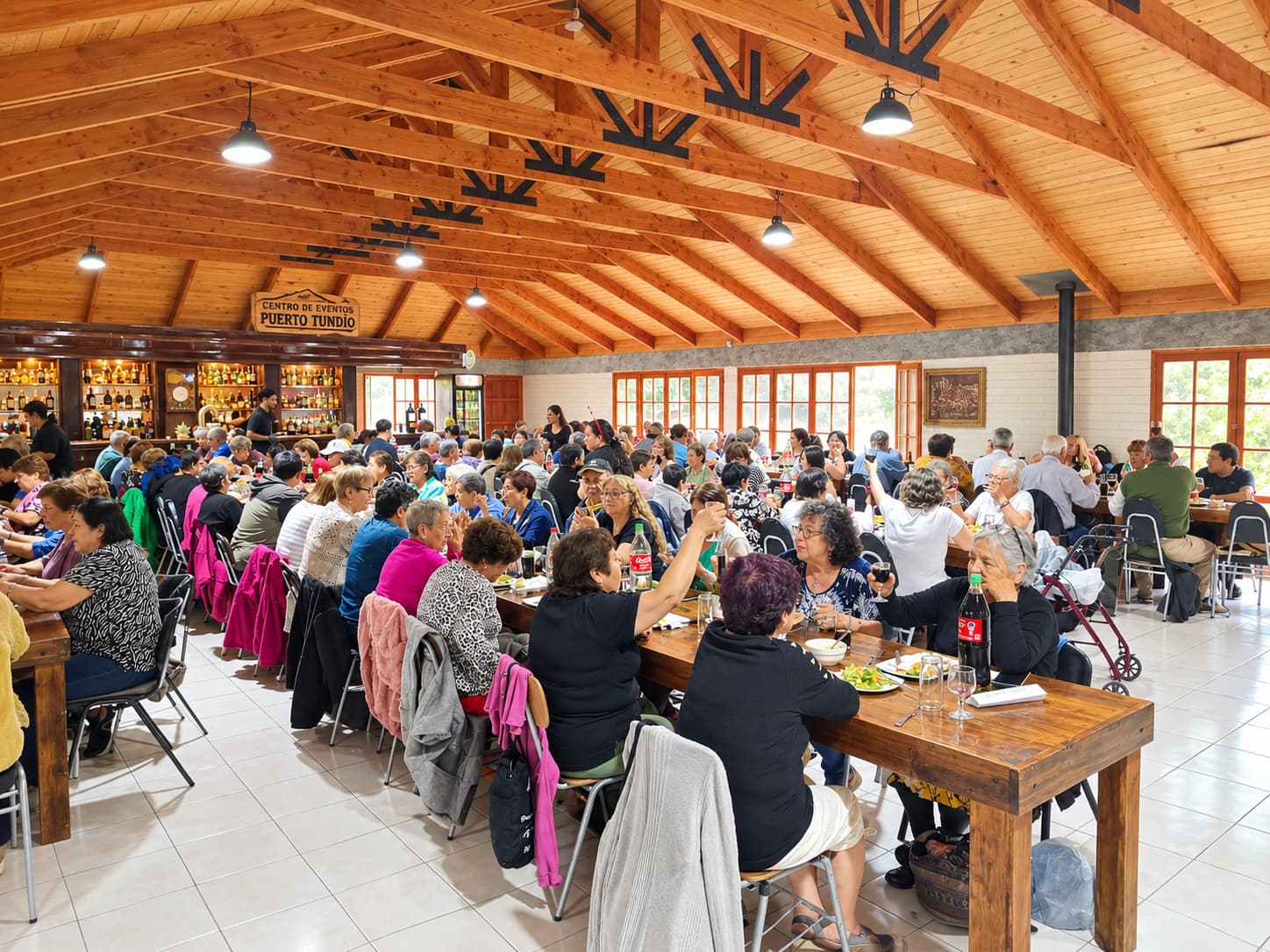 Salón de Pueblo Hundio lleno de personas almorzando y brindando en mesas largas de madera, bajo el techo de vigas