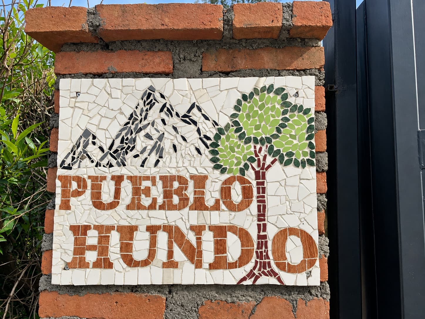 Letrero de mosaico de Pueblo Hundio, con montañas y un árbol, sobre un pilar de ladrillo
